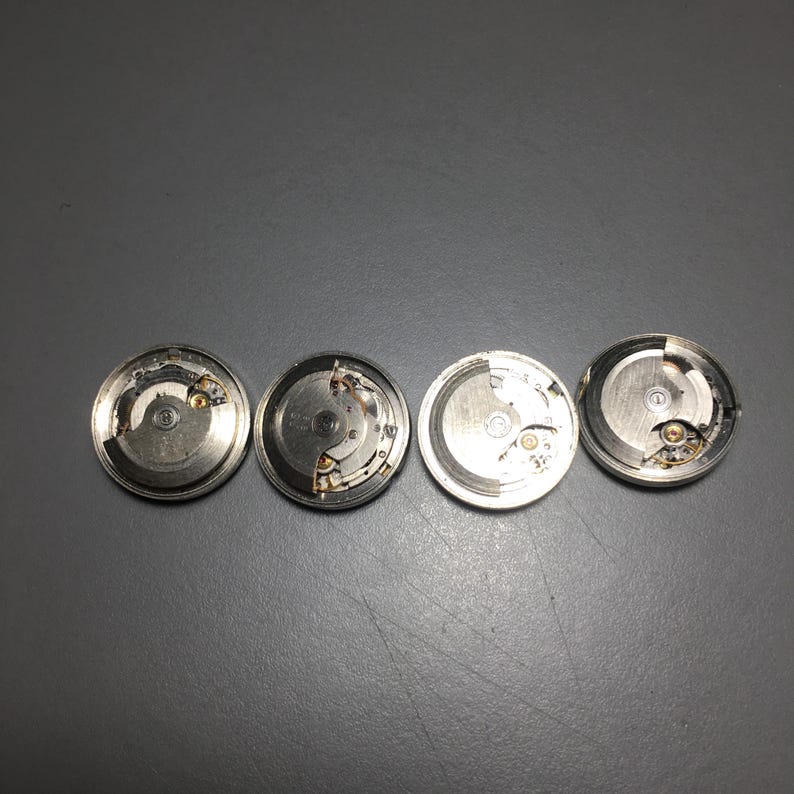 Puede incluir: Se muestran cuatro movimientos de reloj plateados con intrincados mecanismos internos. Las piezas del reloj, de forma circular, revelan engranajes, muelles y otros componentes. La imagen es desde arriba, mostrando la detallada artesan&iacute;a.