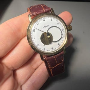 May include: A gold-toned wristwatch with a white face and black numerals. The watch features a unique open design and a brown leather band. The word "PAKETA" is printed on the watch face.