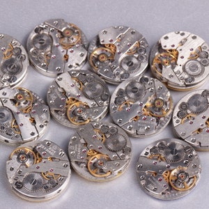 May include: A collection of 10 silver-toned watch movements with intricate gold gears and ruby jewels. The watch movements are arranged in a circular pattern on a silver background.