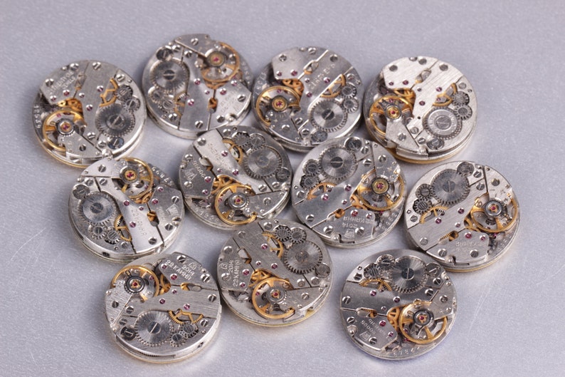 May include: A collection of 10 silver-toned watch movements with intricate gold gears and ruby jewels. The watch movements are arranged in a circular pattern on a silver background.