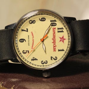May include: A vintage wristwatch with a cream-colored face, black leather band, and silver-tone metal case. The watch face features black numerals, red star, and Cyrillic text. The watch is resting on a brown surface.