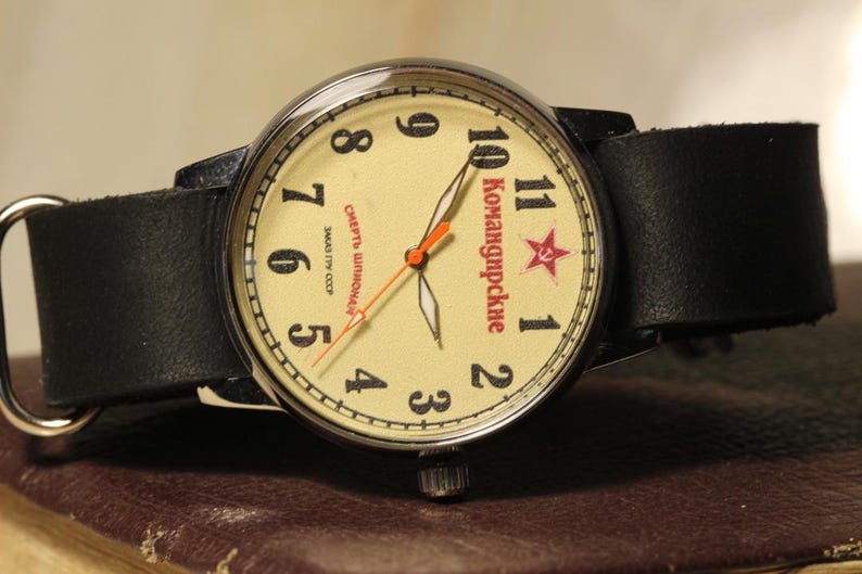 May include: A vintage wristwatch with a cream-colored face, black leather band, and silver-tone metal case. The watch face features black numerals, red star, and Cyrillic text. The watch is resting on a brown surface.