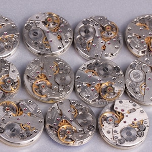 May include: Twelve silver-toned watch movements with intricate gears and red jewels. The movements are arranged in a circle on a gray surface.