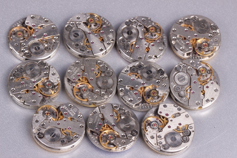 May include: Twelve silver-toned watch movements with intricate gears and red jewels. The movements are arranged in a circle on a gray surface.