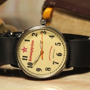 May include: A vintage wristwatch with a cream-colored face, black numerals, and a red star. The watch has a black leather band and silver-colored metal casing. The watch face has Cyrillic text. The watch is set against a blurred background.