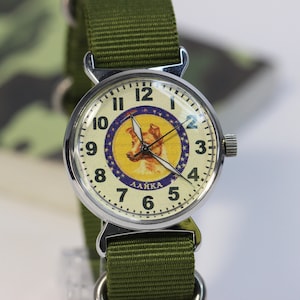 May include: A silver wristwatch with a white face and black numbers. The watch face features a cartoon image of a dog with the word "LAIKA" printed below. The watch has a green canvas strap.