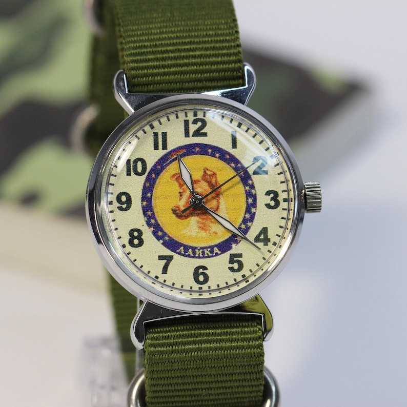 May include: A silver wristwatch with a white face and black numbers. The watch face features a cartoon image of a dog with the word "LAIKA" printed below. The watch has a green canvas strap.