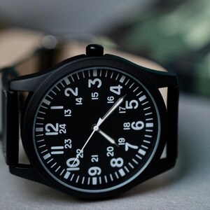 May include: A black wristwatch with a black face and white numerals. The watch has a black case and a leather band. The watch face displays the numbers 1 through 24. The watch hands are white.