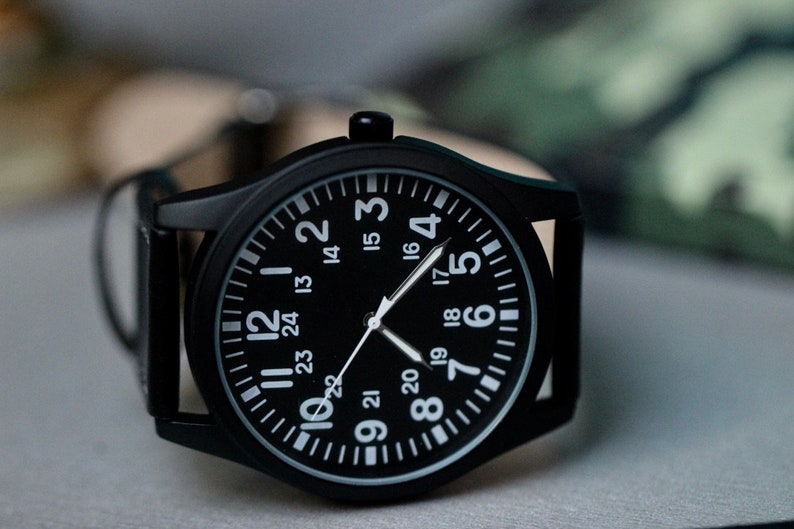 May include: A black wristwatch with a black face and white numerals. The watch has a black case and a leather band. The watch face displays the numbers 1 through 24. The watch hands are white.