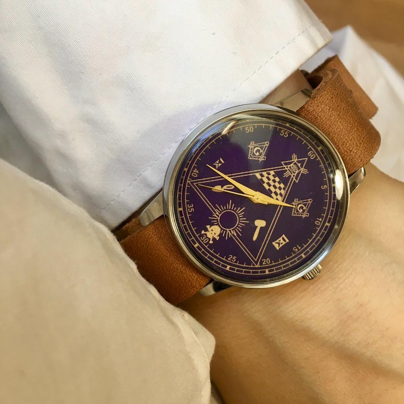 May include: A wristwatch with a dark purple face featuring gold-colored Masonic symbols and hands. The watch has a silver-colored case and a brown leather band. The watch is being worn on a wrist.
