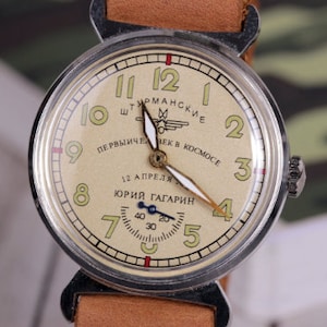 May include: A silver wristwatch with a cream-colored face. The face has black Roman numeral hour markers and black Arabic numeral minute markers. The watch has a brown leather band and the text "ШТУРМАНСКИЕ ПЕРВЫЙЧЕЛ ВЕК В КОСМOCE 12 АПРЕЛЯ ЮРИЙ ГАГАРИН" printed on the face.