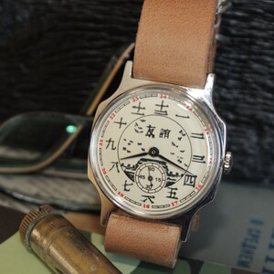 May include: A vintage wristwatch with a cream-colored face featuring Chinese characters and a small illustration. The watch has a silver case and a light brown leather strap. The watch is displayed with eyeglasses and a camouflage pattern.