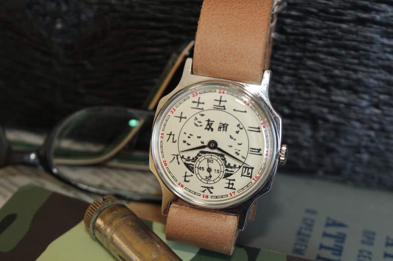 May include: A vintage wristwatch with a cream-colored face featuring Chinese characters and a small illustration. The watch has a silver case and a light brown leather strap. The watch is displayed with eyeglasses and a camouflage pattern.