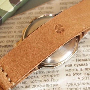 May include: Close-up of a vintage watch with a light brown leather strap. The strap features a stitched design and a stamped cross emblem. The watch face is visible, and the background includes newspaper text.