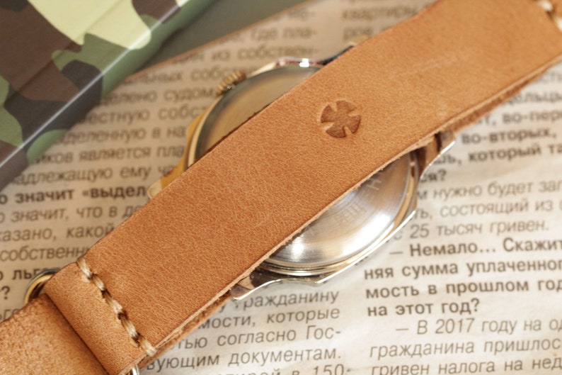 May include: Close-up of a vintage watch with a light brown leather strap. The strap features a stitched design and a stamped cross emblem. The watch face is visible, and the background includes newspaper text.