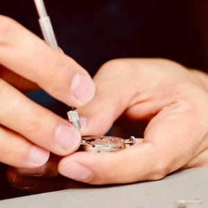 May include: A close-up of a person's hands working on a silver watch mechanism with a small screwdriver.