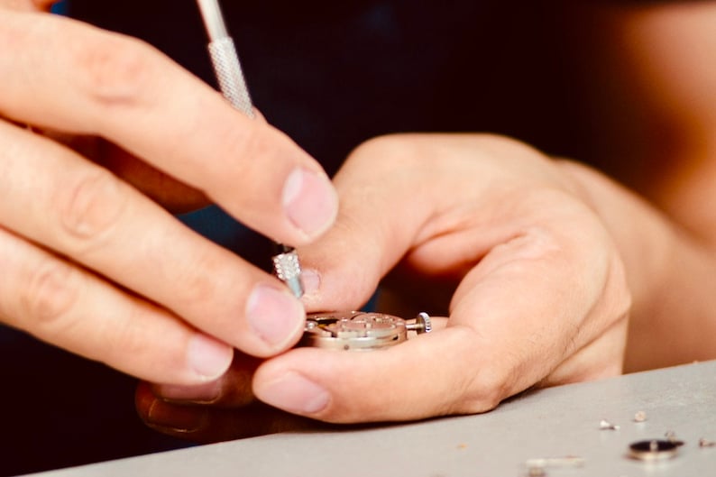 May include: A close-up of a person's hands working on a silver watch mechanism with a small screwdriver.