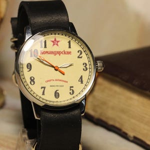 May include: A vintage wristwatch with a cream-colored face, black leather band, and silver casing. The watch face features red numerals, a red star, and the text "Komandirskie". The hands are orange. The watch is set against a blurred background.