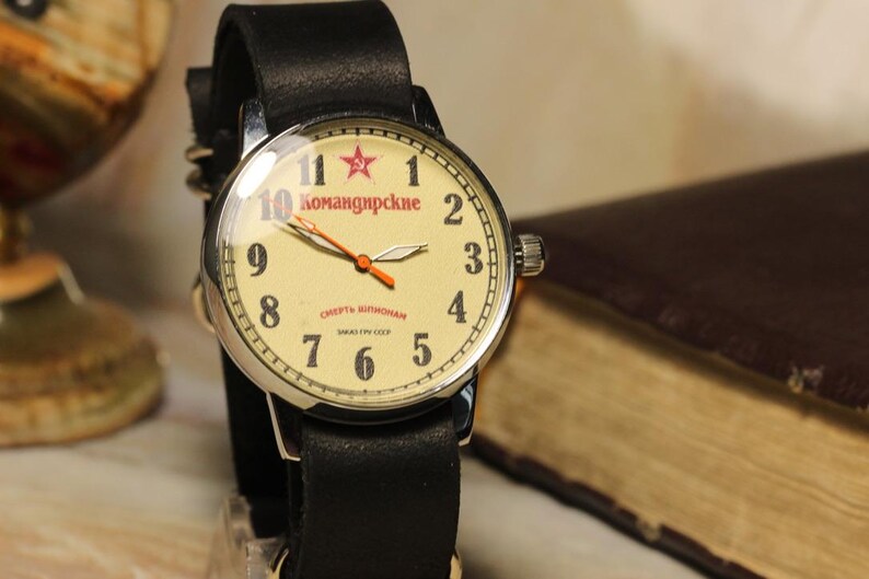 May include: A vintage wristwatch with a cream-colored face, black leather band, and silver casing. The watch face features red numerals, a red star, and the text "Komandirskie". The hands are orange. The watch is set against a blurred background.