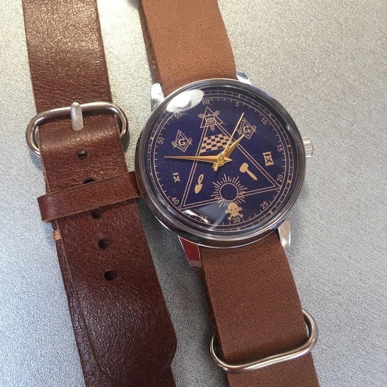 May include: A wristwatch with a dark blue face featuring gold-colored Masonic symbols and hands. The watch has a silver-colored case and a brown leather strap. The strap is connected to the watch with a silver-colored metal bar.