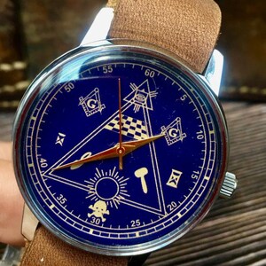 May include: A silver-cased wristwatch with a blue face featuring Masonic symbols in gold. The watch has gold hands and a brown leather strap. The watch face includes a triangle, square, and compass design.