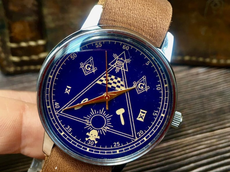May include: A silver-cased wristwatch with a blue face featuring Masonic symbols in gold. The watch has gold hands and a brown leather strap. The watch face includes a triangle, square, and compass design.