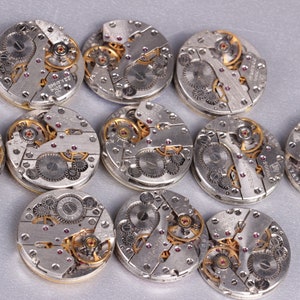 May include: Twelve silver and gold watch movements with intricate gears and mechanisms. The watch movements are arranged in a circle on a gray surface.