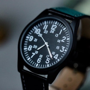 May include: Black wristwatch with a round face and white numerals. The watch has a black case and a black leather strap with white stitching. The watch face displays the numbers 1 through 24. The watch hands are silver.