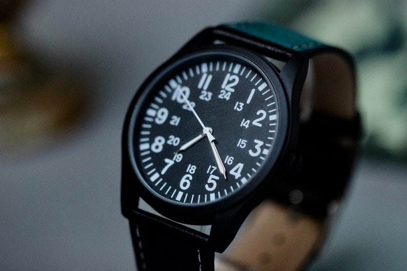 May include: Black wristwatch with a round face and white numerals. The watch has a black case and a black leather strap with white stitching. The watch face displays the numbers 1 through 24. The watch hands are silver.