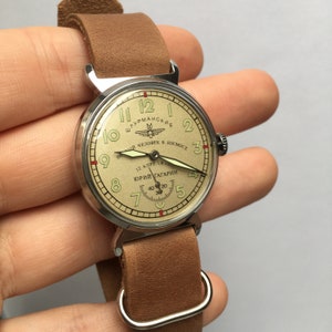 May include: A vintage wristwatch with a round, silver-tone case and a brown leather strap. The watch face is cream-colored with black numerals and hands. The watch face also features the text "Штурманские" and "Первый человек в космосе" in Cyrillic script, along with a small image of a rocket ship. The watch face also features the text "12 апреля 1961" and "Юрий Гагарин" in Cyrillic script.