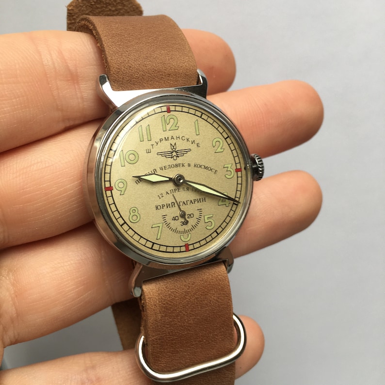 May include: A vintage wristwatch with a round, silver-tone case and a brown leather strap. The watch face is cream-colored with black numerals and hands. The watch face also features the text "Штурманские" and "Первый человек в космосе" in Cyrillic script, along with a small image of a rocket ship. The watch face also features the text "12 апреля 1961" and "Юрий Гагарин" in Cyrillic script.