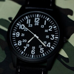 May include: Black wristwatch with a black face and white numerals. The watch has a black band and is set against a camouflage pattern background. The watch face displays both 12-hour and 24-hour time.