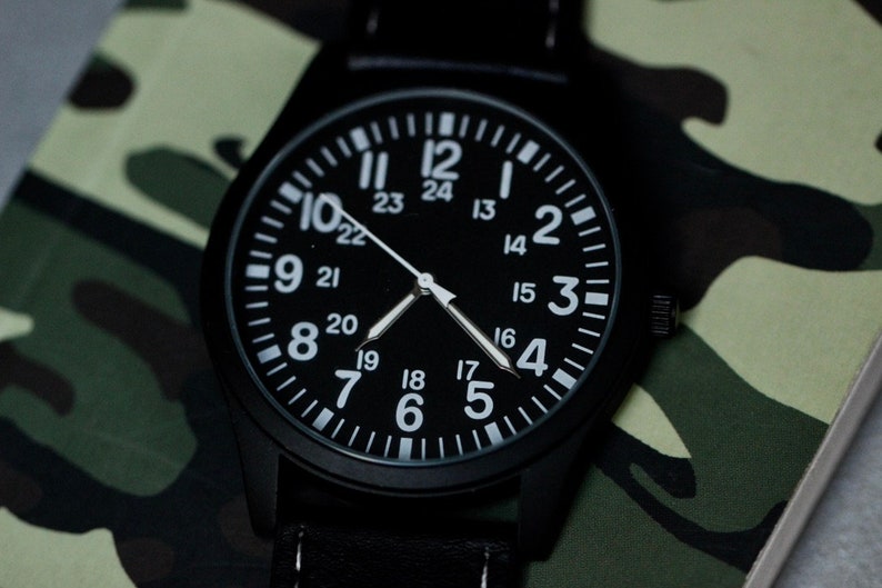 May include: Black wristwatch with a black face and white numerals. The watch has a black band and is set against a camouflage pattern background. The watch face displays both 12-hour and 24-hour time.