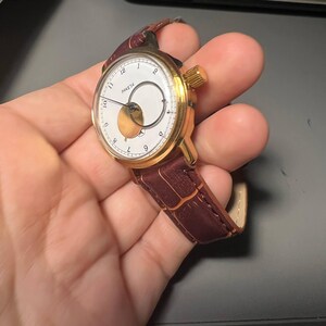 May include: A gold-toned wristwatch with a white face and a brown leather band. The watch face features black numerals and hands, with a unique circular design. The band is a rich, dark brown color.