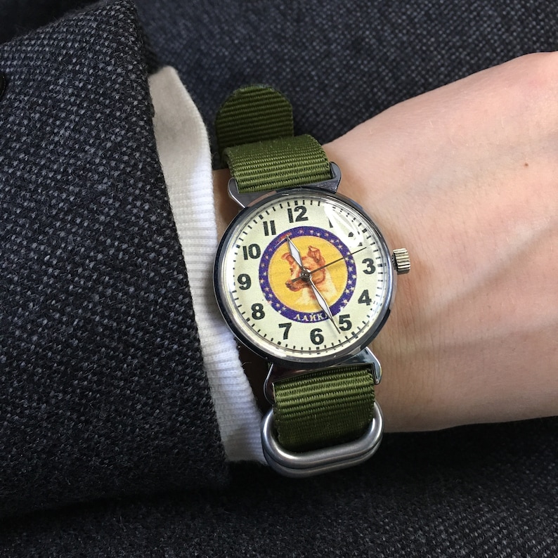 May include: A silver wristwatch with a white face and black numbers. The watch has a green nylon strap and a picture of a dog on the face with the text "LAIKA" underneath.