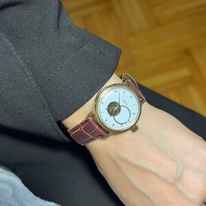 May include: A gold-toned wristwatch with a white face and a brown leather band. The watch features a unique design with a small, circular subdial and a second hand. The band is a deep burgundy color. The watch is a classic accessory.