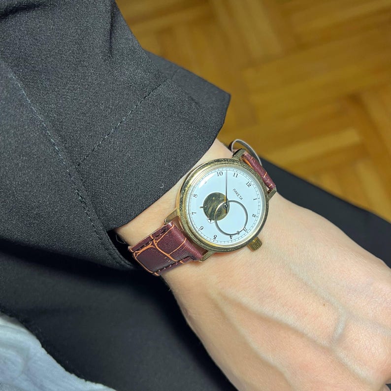 May include: A gold-toned wristwatch with a white face and a brown leather band. The watch features a unique design with a small, circular subdial and a second hand. The band is a deep burgundy color. The watch is a classic accessory.