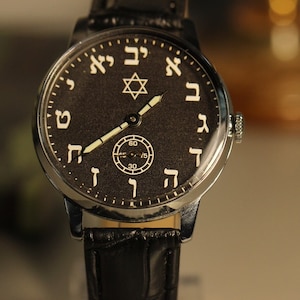 Könnte beinhalten: Eine silberne Armbanduhr mit schwarzem Zifferblatt und hebräischen Ziffern. Die Uhr hat ein schwarzes Lederarmband und einen Davidstern oben auf dem Zifferblatt.