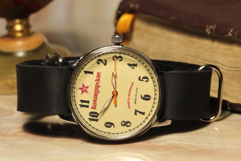 May include: A vintage wristwatch with a cream-colored face, black numerals, and a red star. The watch has a black leather band and silver-colored metal casing. The watch face has Cyrillic text. The watch is set against a blurred background.