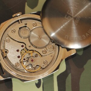 May include: An open-faced, vintage wristwatch showcasing its intricate internal mechanisms. The silver-toned watch reveals gears, springs, and jewels, with the back cover removed. The background is a camouflage pattern.