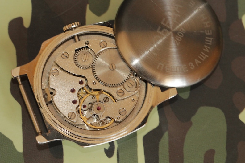 May include: An open-faced, vintage wristwatch showcasing its intricate internal mechanisms. The silver-toned watch reveals gears, springs, and jewels, with the back cover removed. The background is a camouflage pattern.