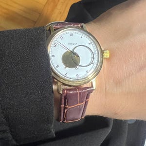 May include: A gold-toned wristwatch with a white face and a brown leather band. The watch face features black numerals and the brand name "PAKETA". The band is a deep burgundy color.
