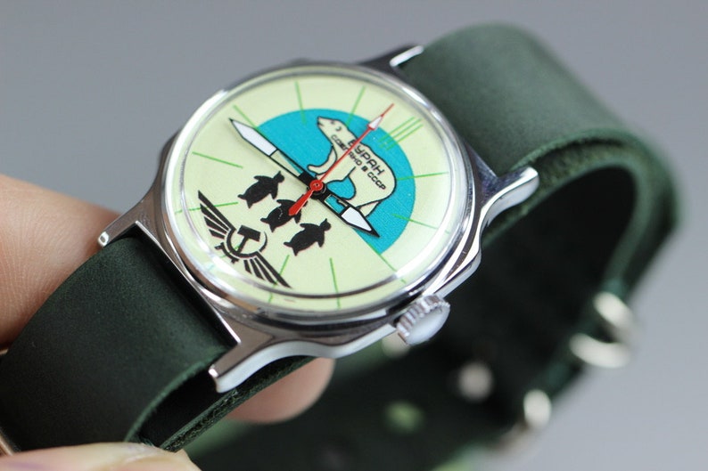 May include: A vintage wristwatch with a green leather band. The watch face features a white background with a green and blue design of a polar bear and penguins. The text "СССР" is printed on the watch face.