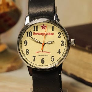 May include: A vintage-style wristwatch with a cream-colored face, black numerals, and a red star at the 12 o'clock position. The watch has a black leather band and a silver-colored case. The watch face displays Cyrillic text. The watch is a classic timepiece.