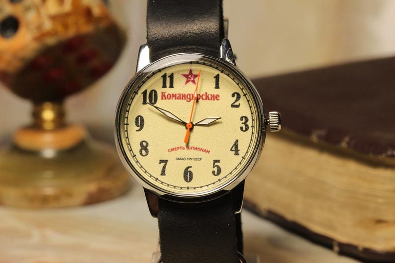 May include: A vintage-style wristwatch with a cream-colored face, black numerals, and a red star at the 12 o'clock position. The watch has a black leather band and a silver-colored case. The watch face displays Cyrillic text. The watch is a classic timepiece.