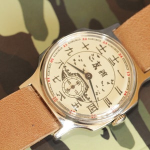 May include: Vintage wristwatch with a cream-colored face featuring Chinese characters and a small seconds dial. The watch has a silver-toned case and a light brown leather strap. The background is a camouflage pattern.