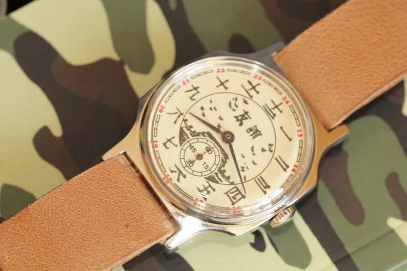 May include: Vintage wristwatch with a cream-colored face featuring Chinese characters and a small seconds dial. The watch has a silver-toned case and a light brown leather strap. The background is a camouflage pattern.
