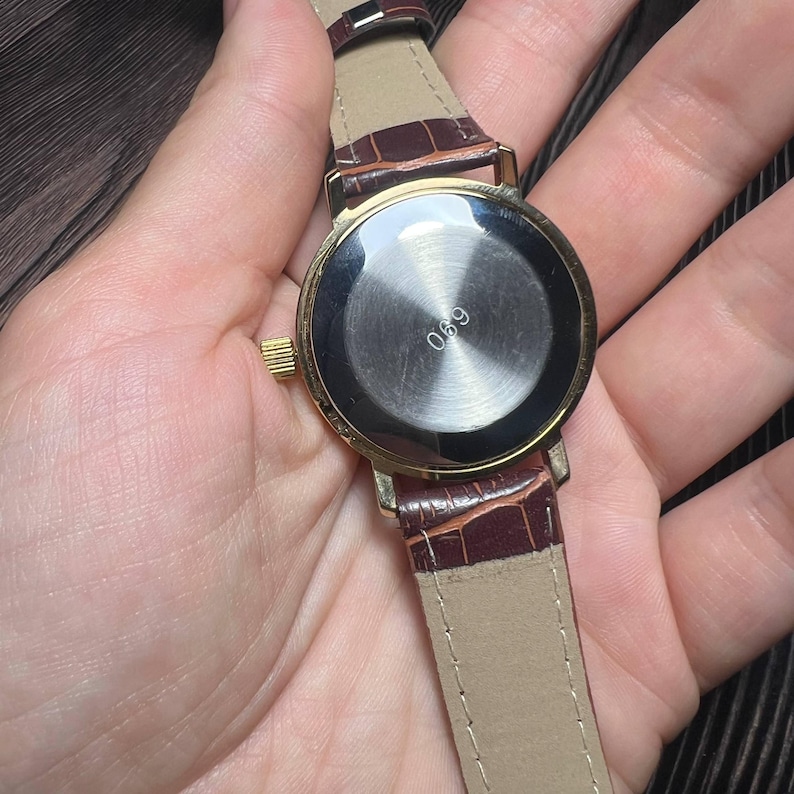 May include: A gold-toned wristwatch with a brown leather band. The watch face is round with a silver-colored center and the number "690" is visible. The band has a crocodile pattern.