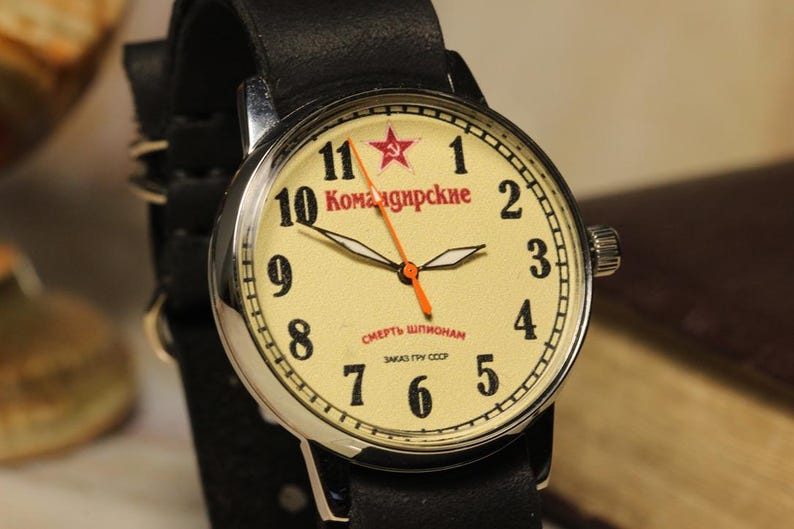 May include: A vintage wristwatch with a cream-colored face, black numerals, and red text. The watch features a red star and an orange second hand. It has a black leather strap and a silver-colored case.