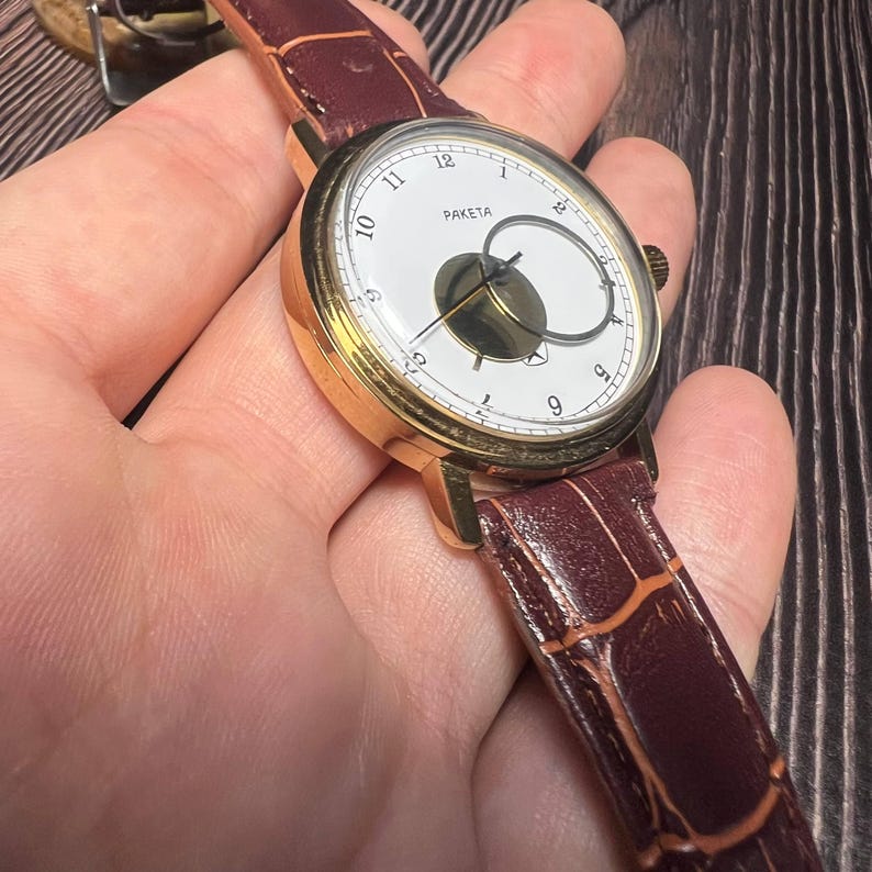 May include: A gold-toned wristwatch with a white face and a brown leather band. The watch face features black numerals and the word "PAKETA". The band is a dark brown with a textured pattern.